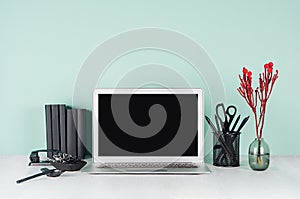 Spring workplace with blank notebook screen, black stationery, books, coffee cup, red flowers in elegant green mint menthe.