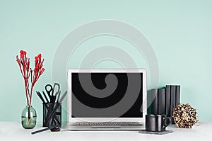 Spring workplace with blank notebook screen, black stationery, books, coffee cup, sheaf of dry branches, red flowers in green mint