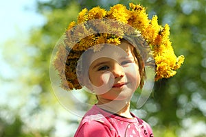 Springtime sunlit portrait of a cute two years old girl posing with a dandelion wreath, looking at the camera