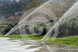 sprinklers water the green lawn in the city park on a summer day, people walking and children playing, bright sunlight