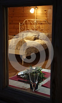 Spy view through window of wooden country house, made bed, set table for romantic dinner, glass of wine, plates and nightlight,