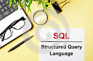 SQL Structured Query Language is written in red on a white piece of paper on a light yellow background next to a laptop, pen,