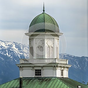 Square Building exterior with green dome and roof against snowy mountain and cloudy sky