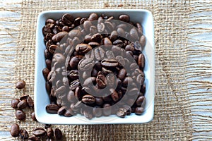 SQUARE CERAMIC BOWL CONTAINING ROASTED COFFEE BEANS WITH LOOSE BEANS ON A PIECE OF HESSIAN