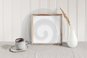 Square empty wooden frame mockup with modern ceramic vase, dry grass, cup of coffee on table. White beadboard wainscot