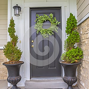 Square Gray front door of a home with green wreath and flanked by tall potted plants