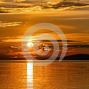 Square photo of Sunset over Umea river Mountains, summer sky with clouds highlighted by orange red Sun. Sunlight path on water