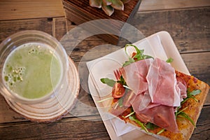 Square pizza slice with prosciutto, arugula and cherry tomatoes green juice in a cup