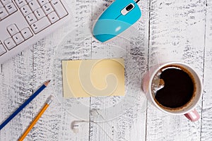 Square rectangle empty paper note above a white wooden floor, computer keyboard and mouse. Mug of coffee and pencils