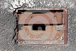 Square rusted iron drain opening with missing grates cover surrounded with cracked asphalt