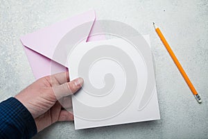 A square sheet of blank white paper in his hand, a pink envelope for mail and a yellow pencil