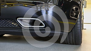 Square tailpipe at the rear right side of a black sports car. Very low plan. The rear bumper, wheel and tailpipe of the packed