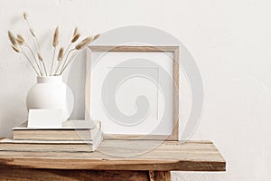 Square wooden frame mockup on vintage bench, table. Modern white ceramic vase with dry Lagurus ovatus grass, books and