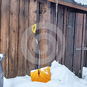 Square Yelllow snow shovel against snowed in ground and brown wooden wall of building