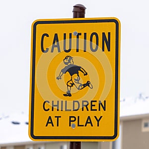 Square Yellow Caution Children At Play sign against blurry building and cloudy sky