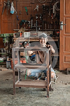 Squat male welder smiles at the camera holding an iron rack to weld