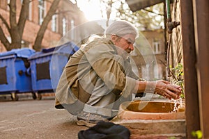 Squating old tramp cleaning face under water.