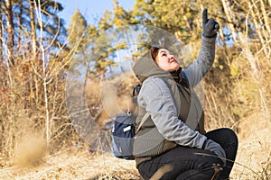 Squatty woman in winter clothes and backpack looking up and raised her hand in gloves showing something.