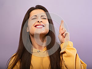 Squint smiling young desiring woman in yellow hoodie crossing fingers with closed eyes on purple bright background. Closeup photo