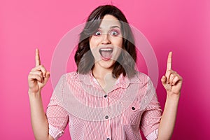Srudio shot of young brunette girl posing isolated over pink background, points fingers up, having great, female wears striped photo