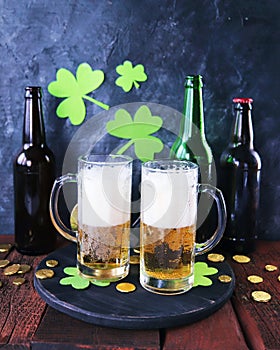 St. Patrick`s Day, fresh beer in glass mugs and a bottle, gold coins, on a red wooden table, a green shamrock on a dark background