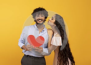 St. Valentine& x27;s day. Pretty indian woman kissing smiling man, holding red heart, yellow background