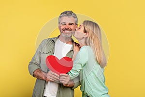 St. Valentines day concept. Loving mature spouses holding red paper heart card, woman kissing her happy husband