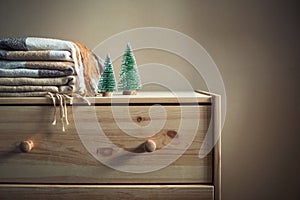 Stack of beige checkered wool blankets and two Christmas trees on a wooden chest. Cozy winter still life. Home textile.