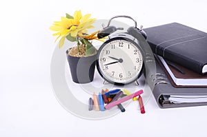 Stack of book with ticking vintage clock, artificial flower plant and colorful crayon on white table.