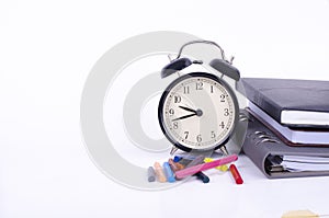 Stack of book with ticking vintage clock, artificial flower plant and colorful crayon on white table.