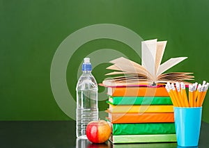 Stack of books with apple, bottle of water and pencils near empty green chalkboard. Sample for text