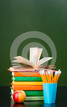 Stack of books with apple and pencils near empty green chalkboard