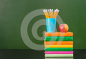 Stack of books with apple and pencils near empty green chalkboard