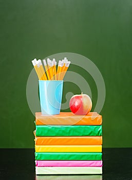 Stack of books with apple and pencils near empty green chalkboard