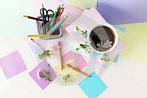 A stack of books, a cup of coffee, pencils, notepads and twigs of cherry blossoms on the table, against a background of colored pa