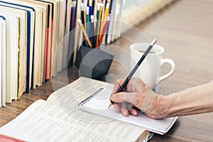 Stack of books education background, female hand makes notes in open notebook. Glasses, pens and pencils in holder, cup of tea