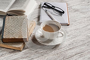 A stack of books, open clean notepad, glasses and a cup of cocoa on a white wooden table.