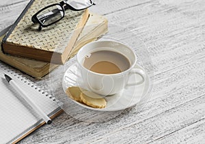 A stack of books, open clean notepad, glasses and a cup of cocoa on a white wooden table.
