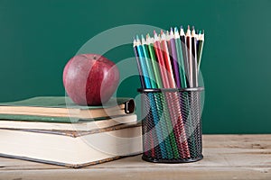 A stack of books with a red apple and a pen holder full of colored pencils in front of the blackboard