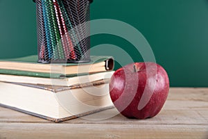 A stack of books with a red apple and a pen holder full of colored pencils in front of the blackboard