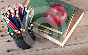 A stack of books with a red apple and a pen holder full of colored pencils in front of the blackboard