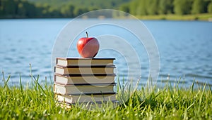 Stack Of Books With Red Apple Placed On Green Grass Beside Peaceful Lake Under Bright Daylight