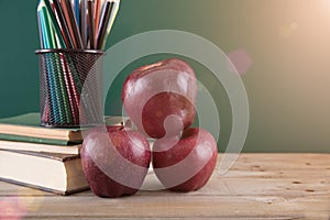 A stack of books and two red apples in front of the blackboard and a pen holder filled with colored pencils