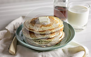 Stack of buttermilk pancakes with butter. Maple syrup and glass of milk in the background.