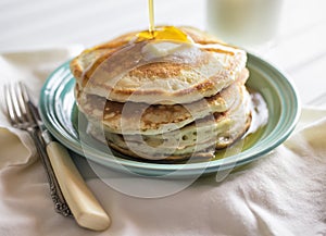 Stack of buttermilk pancakes with butter and syrup being poured on the top. Glass of milk in the background.