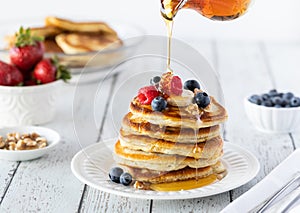 A stack of buttermilk pancakes served with berries and bananas with syrup being poured on top.