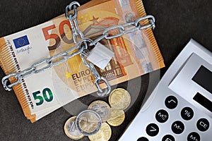A stack of cash with 50 euro banknotes is locked with a chain and padlock next to a calculator and coins