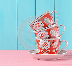 A stack of ceramic retro cups with red patterns on a pink wooden table against a blue wooden wall background, pastel color trend.