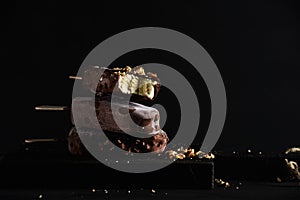 Stack of chocolate dipped popsicles with chipped nuts on dark wooden board over black background.