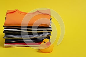 Stack of clean baby clothes and rubber duck on yellow background. Space for text